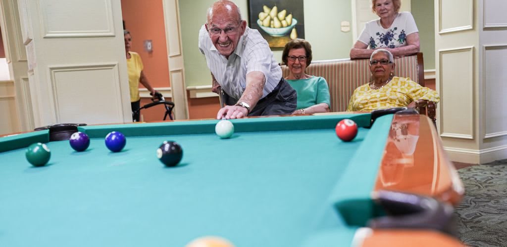 An elderly man playing billiards with friends watching.