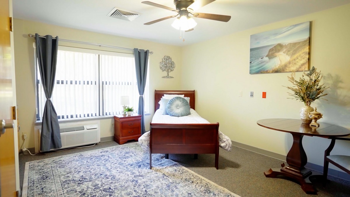 A cozy bedroom with a single bed, nightstand, and ceiling fan, featuring modern decor and natural light from a large window.