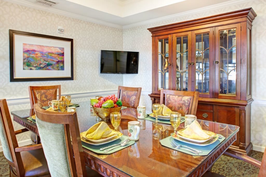 A well-appointed dining room with a polished wooden table, elegantly set with place settings and a fruit bowl, complemented by a china cabinet and wall-mounted television.