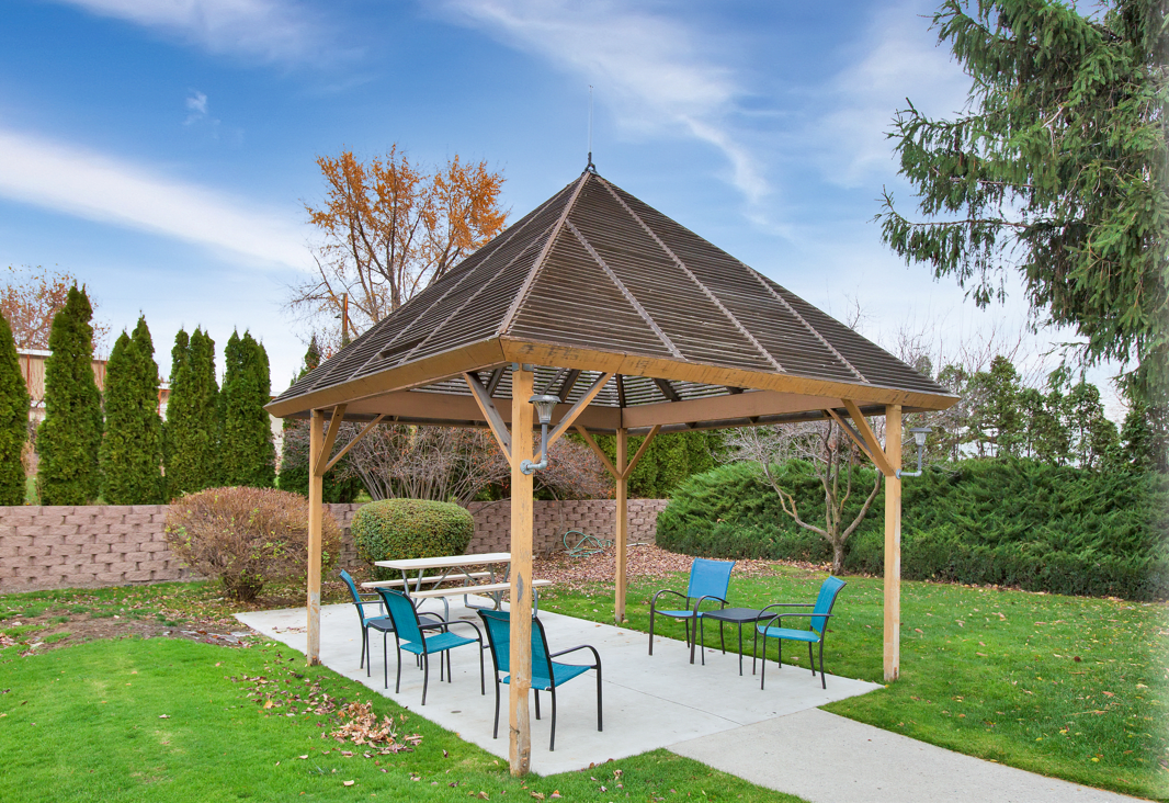 A wooden gazebo with blue chairs and a table, set in a park with green grass and surrounding trees.