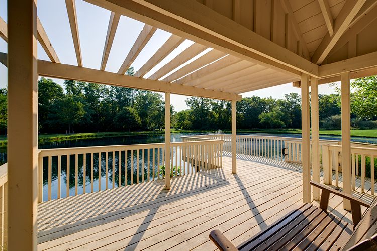 A wooden deck with a pergola overlooking a peaceful lake surrounded by trees.