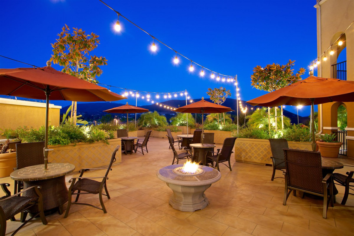 A cozy outdoor patio with string lights and a fire pit, featuring tables and chairs under umbrellas during the evening.