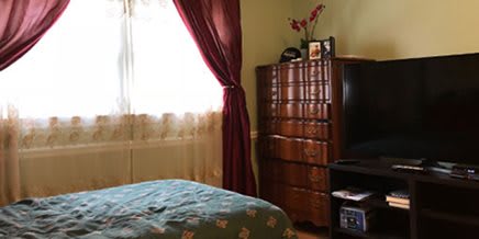A cozy bedroom featuring a window with red curtains, wooden dresser, television, and decorated bed.