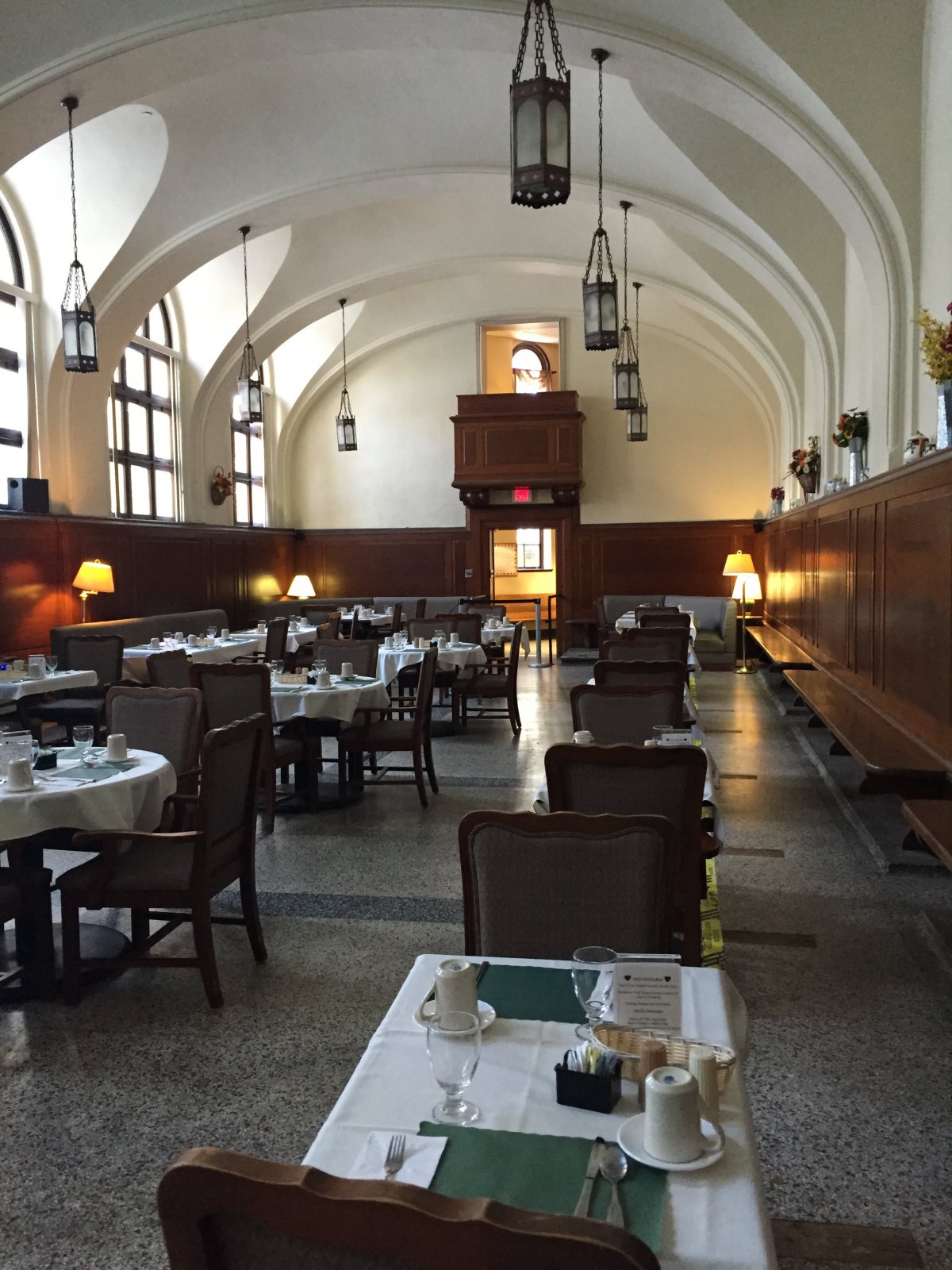 A spacious dining hall with elegantly set tables and an arched ceiling, softly lit by hanging lamps.