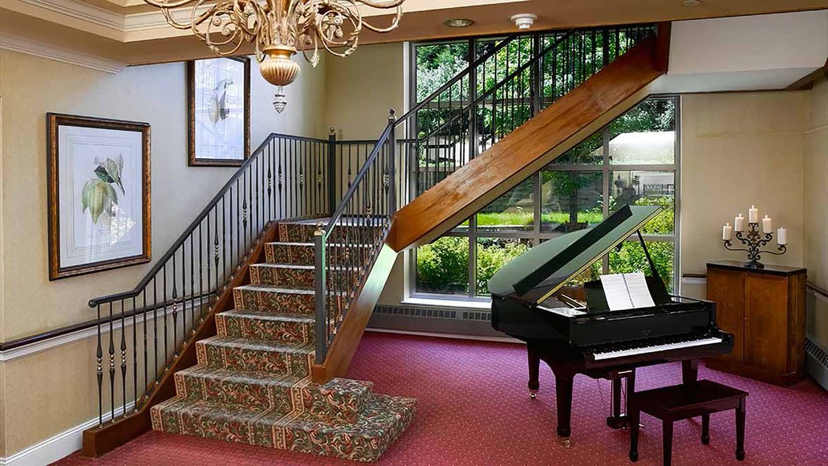 A luxurious interior featuring a grand staircase with a patterned carpet, a grand piano by the window, and an elegant chandelier overhead.