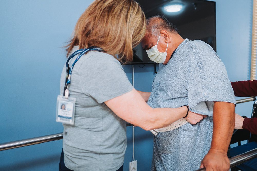 A nurse assists a patient in a hospital setting, both wearing masks.