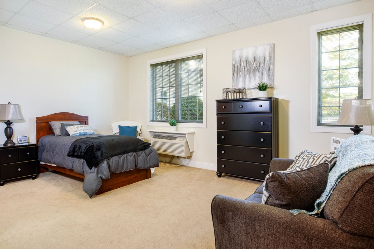 A cozy bedroom with a bed, dresser, and chair near a window.