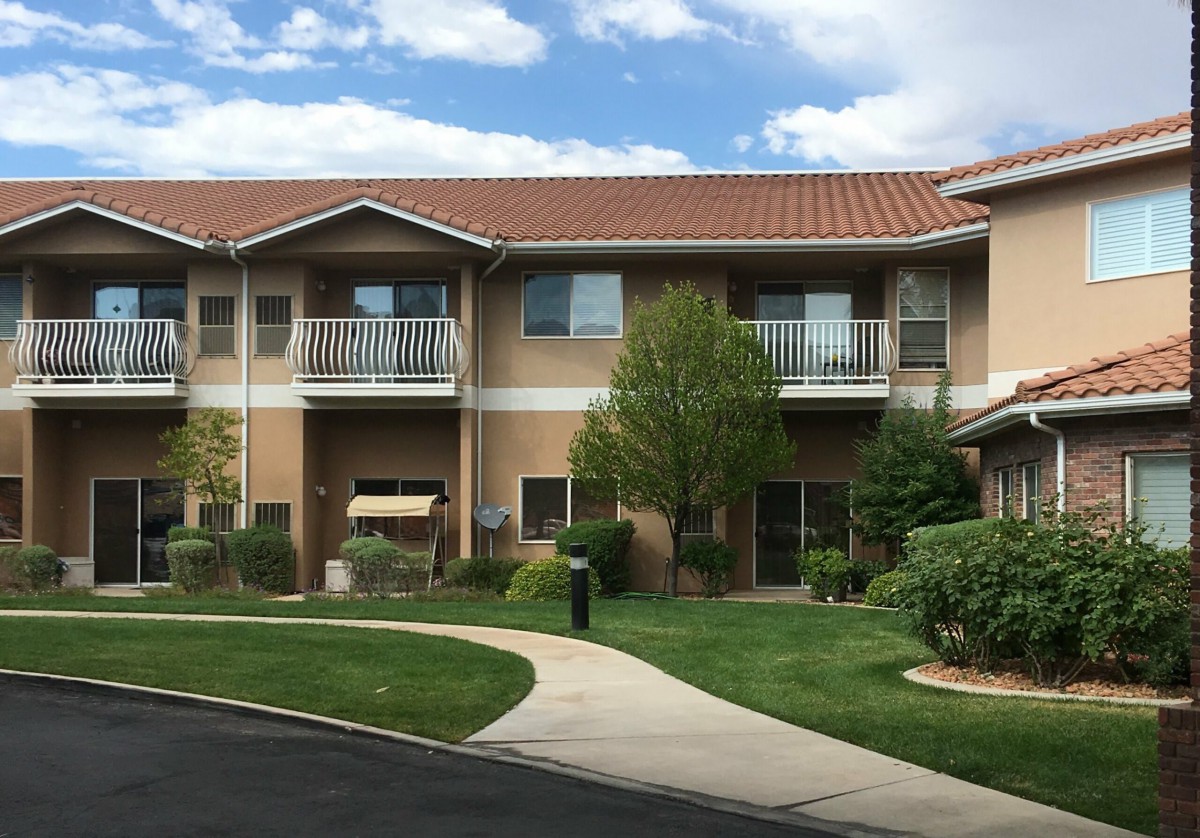 A two-story residential apartment building with a landscaped yard, featuring trees and a grassy area under a partly cloudy sky.