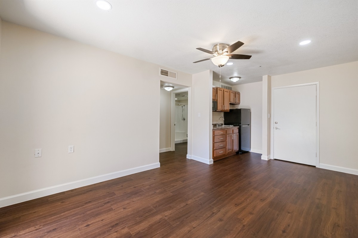 An unfurnished apartment with a living area and kitchenette, featuring hardwood floors and a ceiling fan.