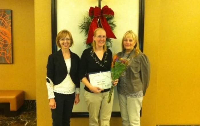 Three individuals standing together, with one person holding a certificate and flowers, celebrating an achievement.