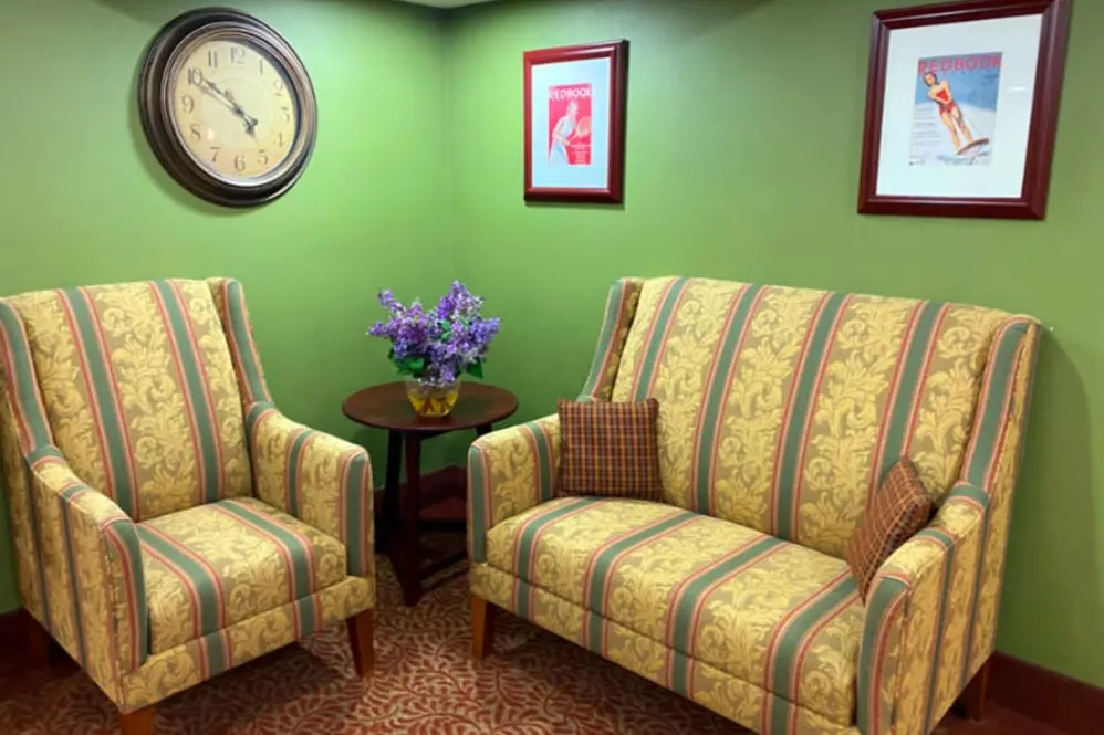 A cozy living room with two patterned sofas, a side table with a flower vase, and artwork on green walls.