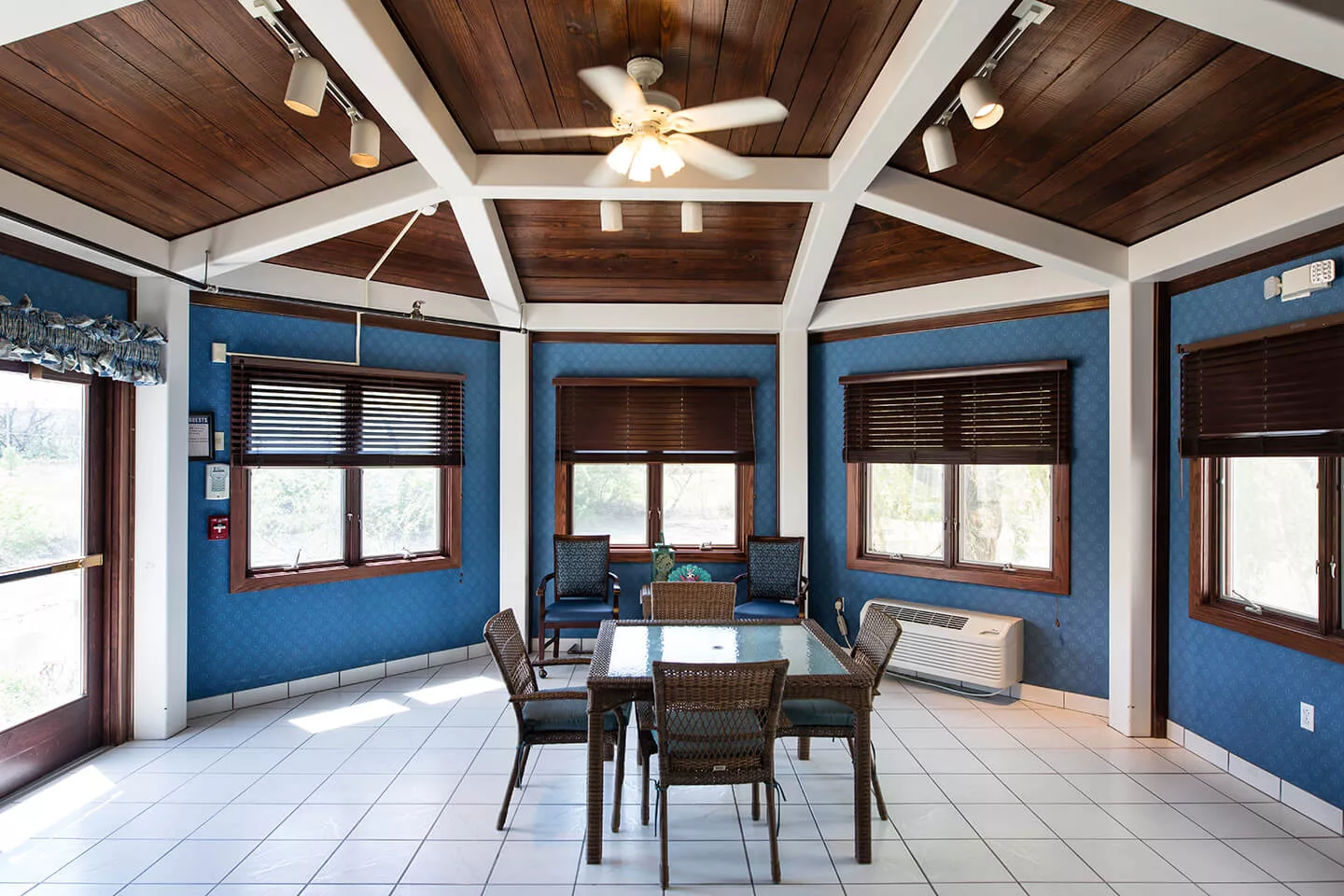 A spacious dining room with a wooden ceiling and large windows, furnished with a glass table and chairs.