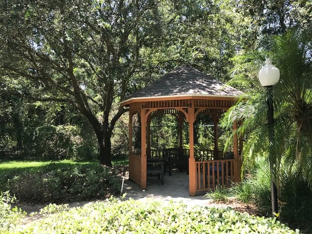 A tranquil garden scene featuring a wooden gazebo surrounded by lush greenery and trees.