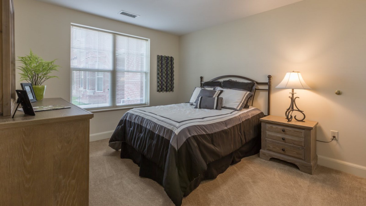 A cozy bedroom featuring a neatly made bed with matching linens, a bedside lamp on a wooden nightstand, and a window allowing natural light to illuminate the room.