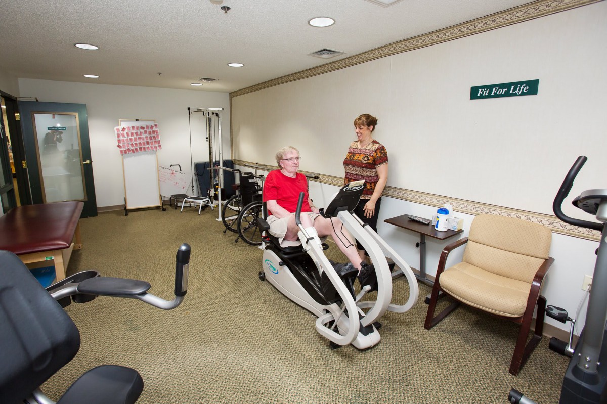 An elderly person using gym equipment for rehabilitation in a healthcare setting with assistance.