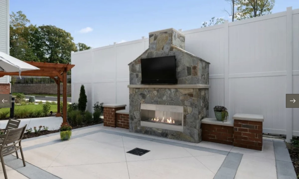 A modern outdoor patio featuring a stone fireplace with a mounted TV and surrounding landscaping.