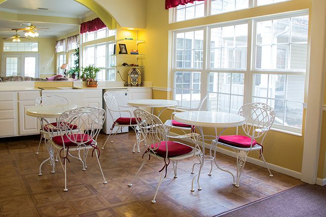 A cozy dining area with white tables and chairs, featuring red cushions and large windows.
