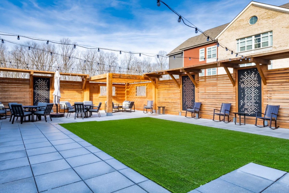 A modern backyard with a patio featuring artificial grass, string lights, and seating under a pergola.