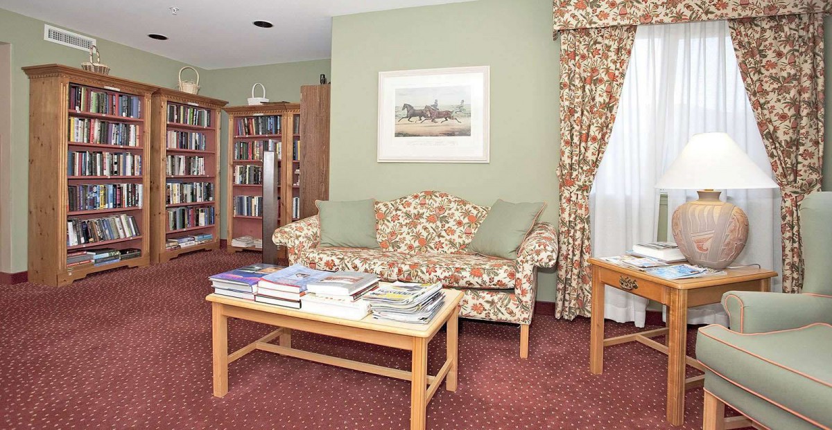 A cozy living room with a floral patterned sofa, a bookshelf filled with books, and a coffee table topped with magazines. The room features green walls, a decorative painting, and matching curtains.