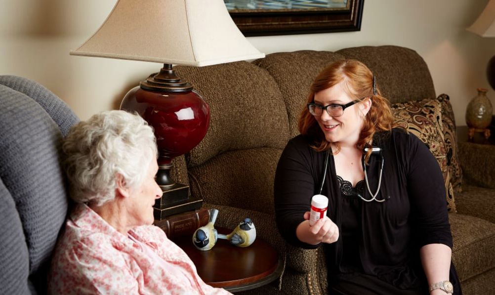 A nurse is talking to an elderly woman during a home visit, holding a bottle of medication.