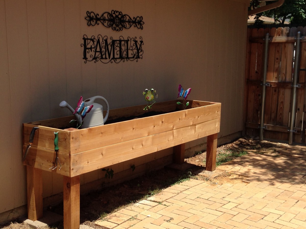 A wooden raised garden bed on a patio, accompanied by watering cans and a 'Family' sign on the wall.