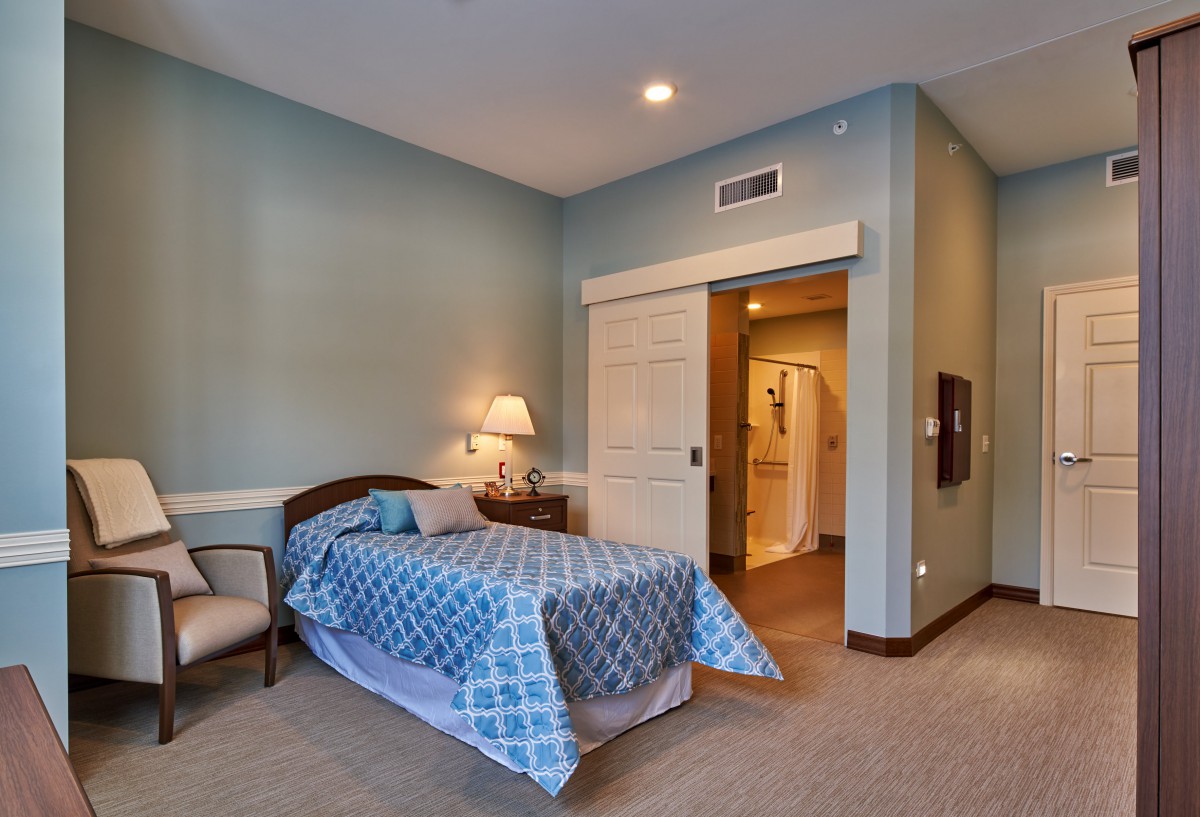 A cozy bedroom with blue walls and a single bed, featuring a lamp, chair, and side table.