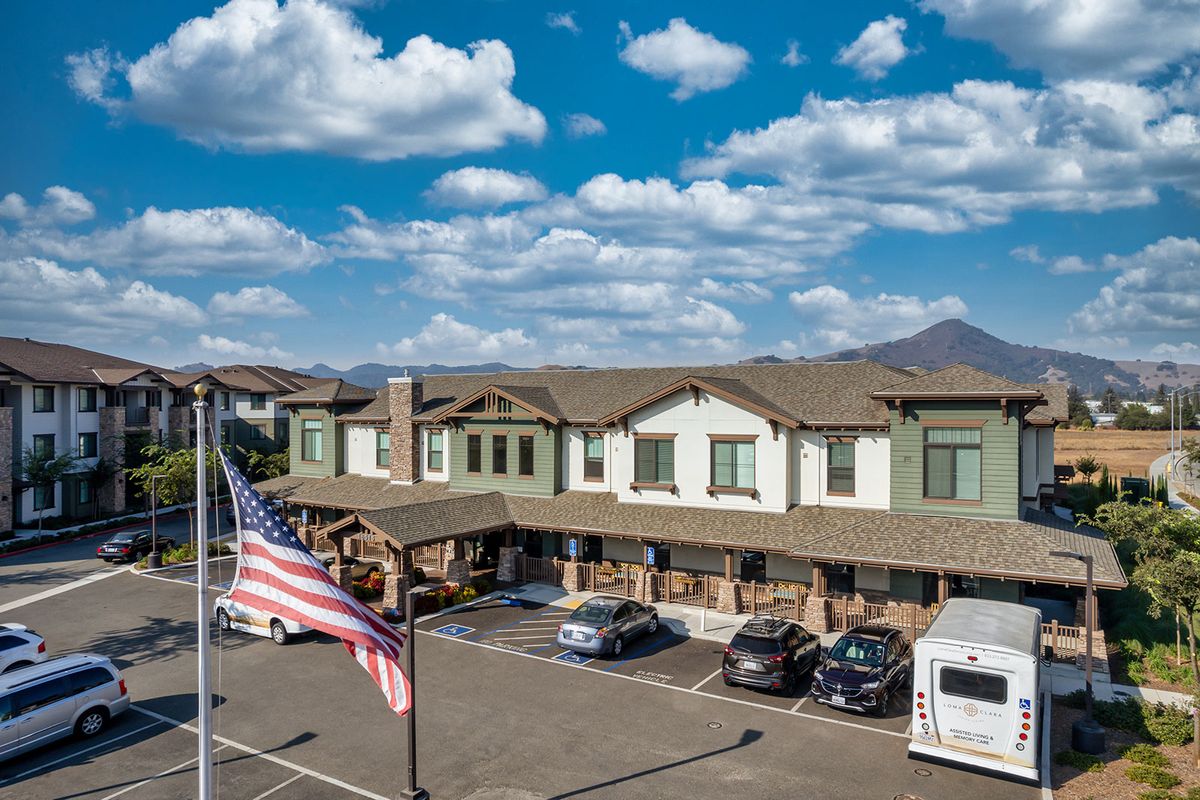 A mid-sized hotel with a parking lot in front, featuring several parked vehicles and an American flag waving under a partly cloudy sky with mountains in the background.