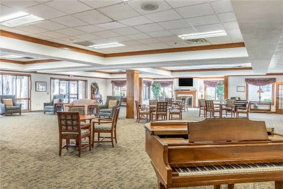 A spacious, well-lit room featuring a piano, several tables and chairs, and large windows.