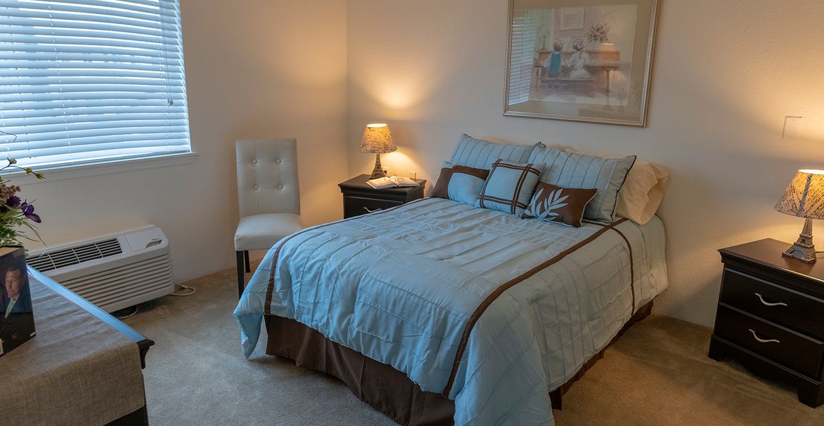 A cozy bedroom with a neatly made bed, a nightstand with a lamp, and a chair in the corner.