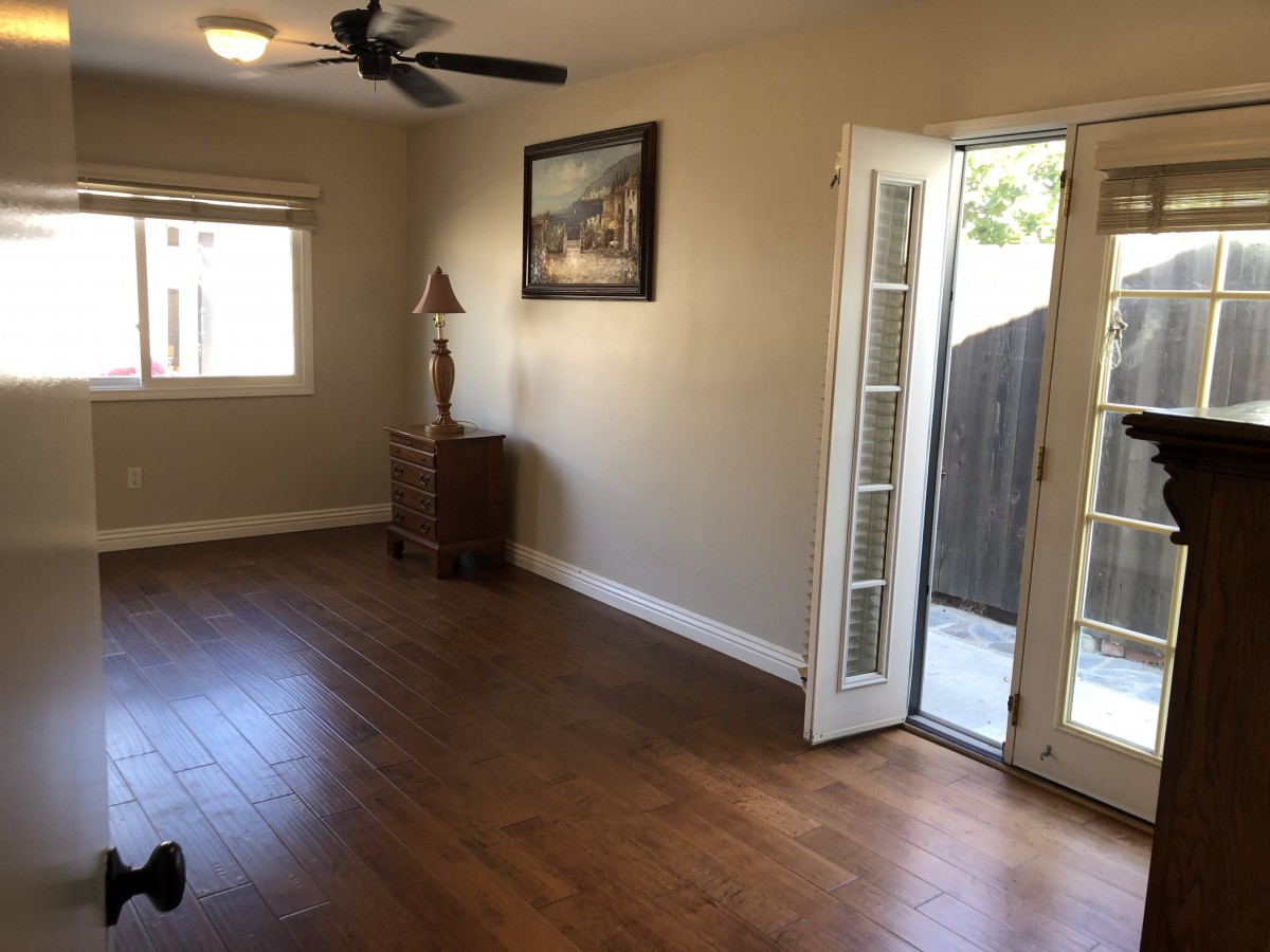 A spacious living room with hardwood floors, featuring a ceiling fan, French doors leading outside, a window, and decorated with a painting and a sculpture.