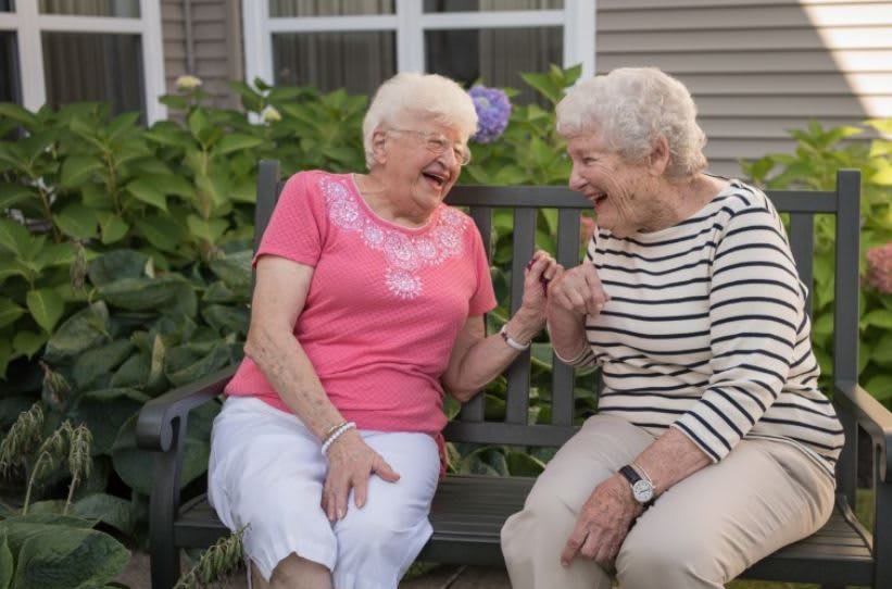Two elderly women share a laugh while sitting on a bench outside.