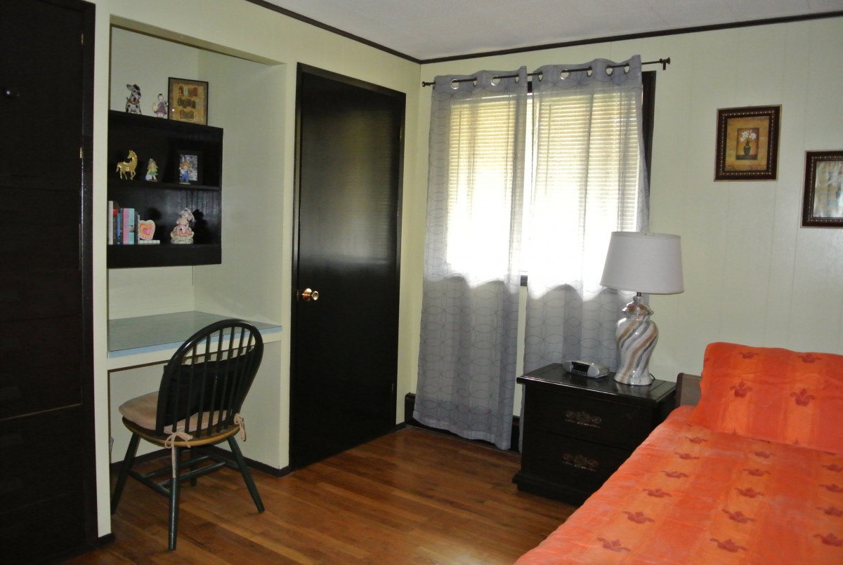 A cozy bedroom with a desk and chair, featuring a window with curtains and a lamp on a side table.