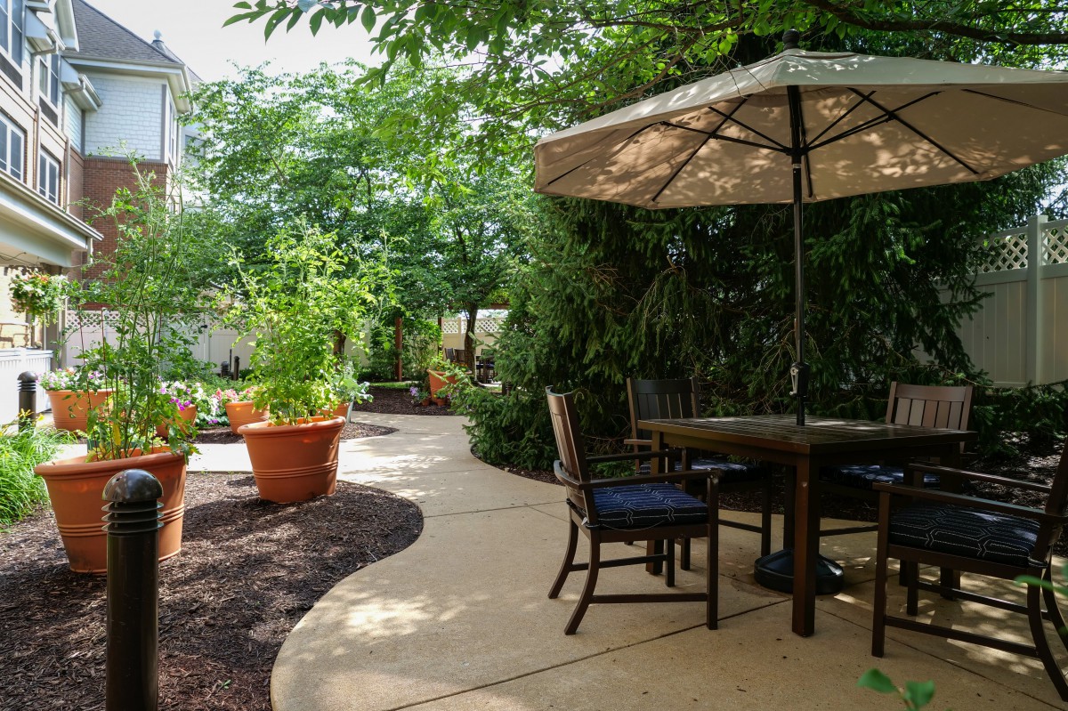 A serene garden patio with a table, chairs, and umbrella surrounded by lush plants.