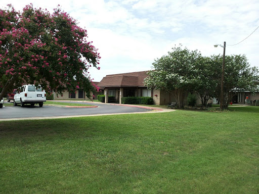 A view of a building surrounded by greenery and a parking area under a cloudy sky.