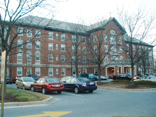 A four-story brick residential building with a parking lot in front, surrounded by trees.