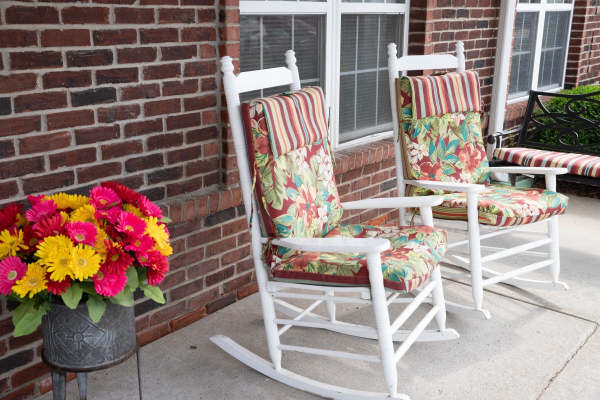 Two white rocking chairs with colorful cushions sit on a porch beside a brick wall and window, accompanied by a pot of bright flowers.