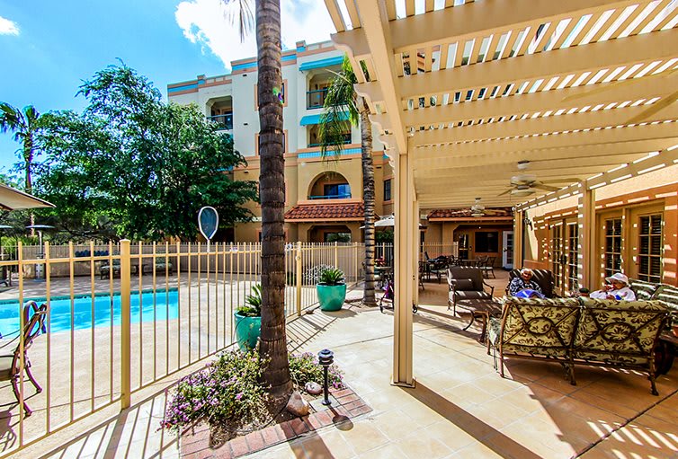 A sunny outdoor patio with a pergola overlooking a pool surrounded by a fence and palm trees.