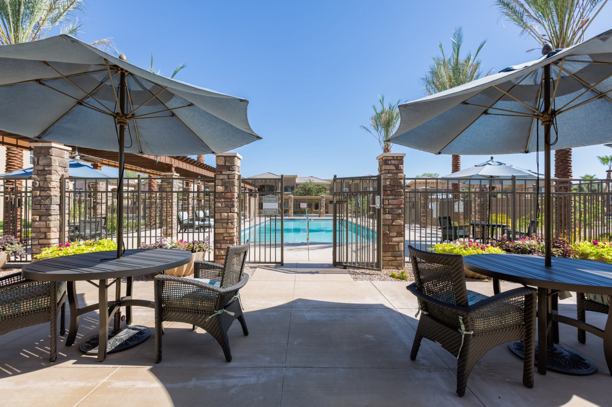 A sunny patio area with tables and umbrellas overlooking a fenced swimming pool.