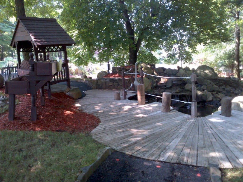 A peaceful garden setting with a wooden walkway leading to a gazebo under lush trees.