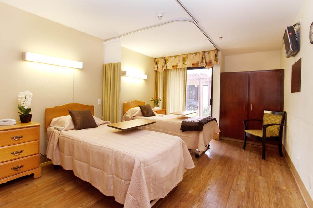 A hospital room with two beds, wooden flooring, and natural light from a window.