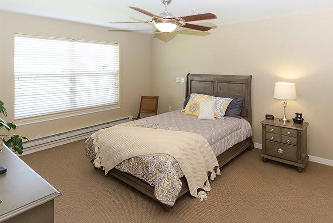 A cozy bedroom with a neatly made bed, ceiling fan, and natural light coming through the window.
