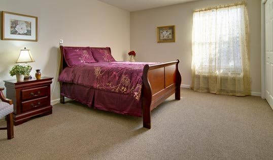 A cozy bedroom with a wooden bed and purple bedding, featuring a large window and carpeted floor.