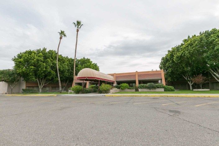 A building surrounded by palm trees and greenery under an overcast sky, with an empty parking lot in the foreground.