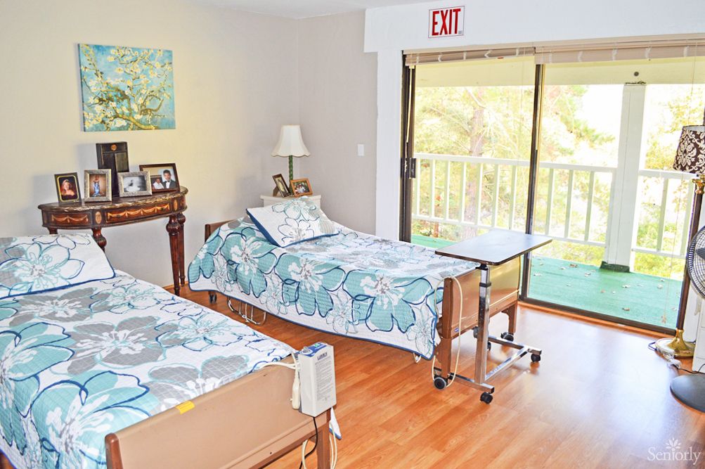 A shared bedroom featuring two hospital beds with floral bedding, a wooden floor, and a balcony view.