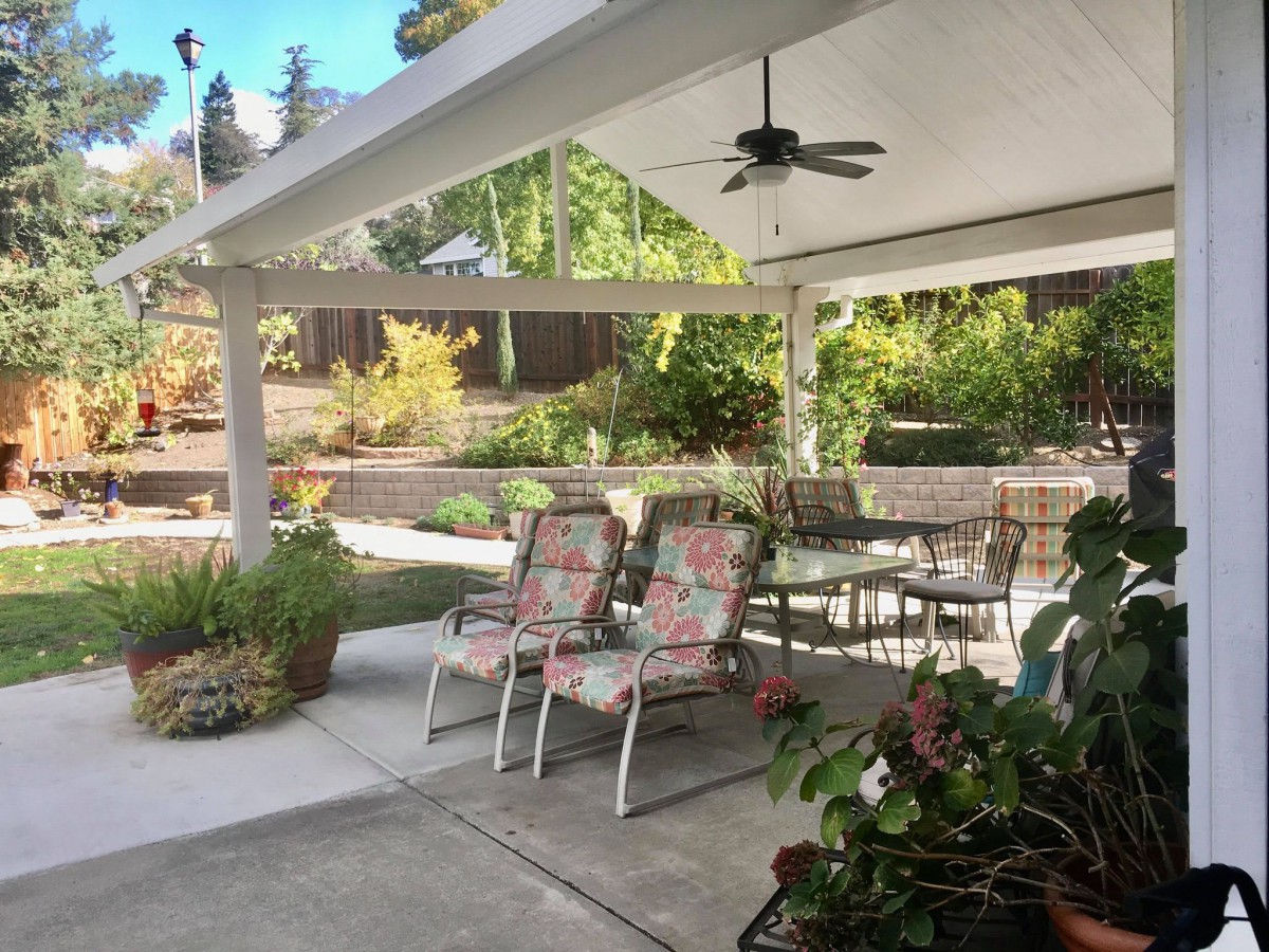 A cozy outdoor patio with chairs and a table under a covered area, surrounded by lush garden plants.