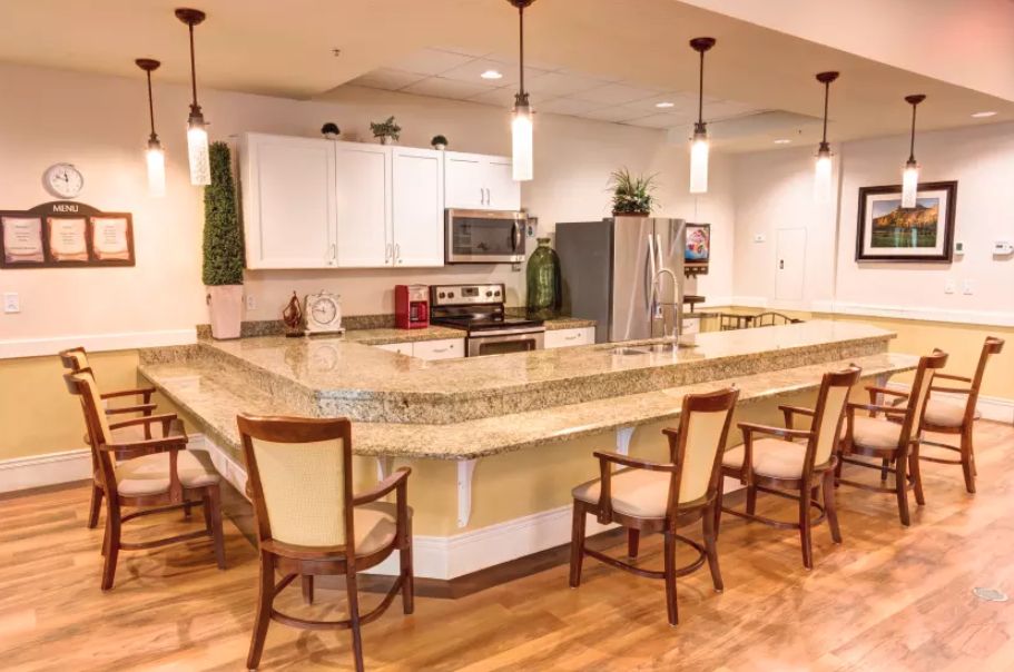 A modern kitchen with a large granite counter and wooden chairs, featuring stainless steel appliances and pendant lighting.
