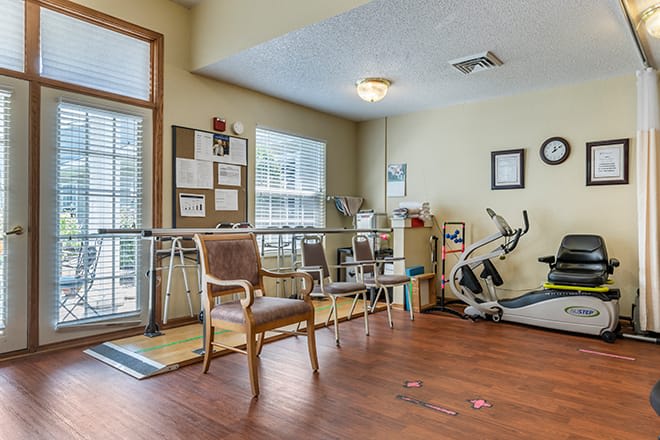 A rehabilitation therapy room featuring exercise equipment and chairs on a wooden floor.