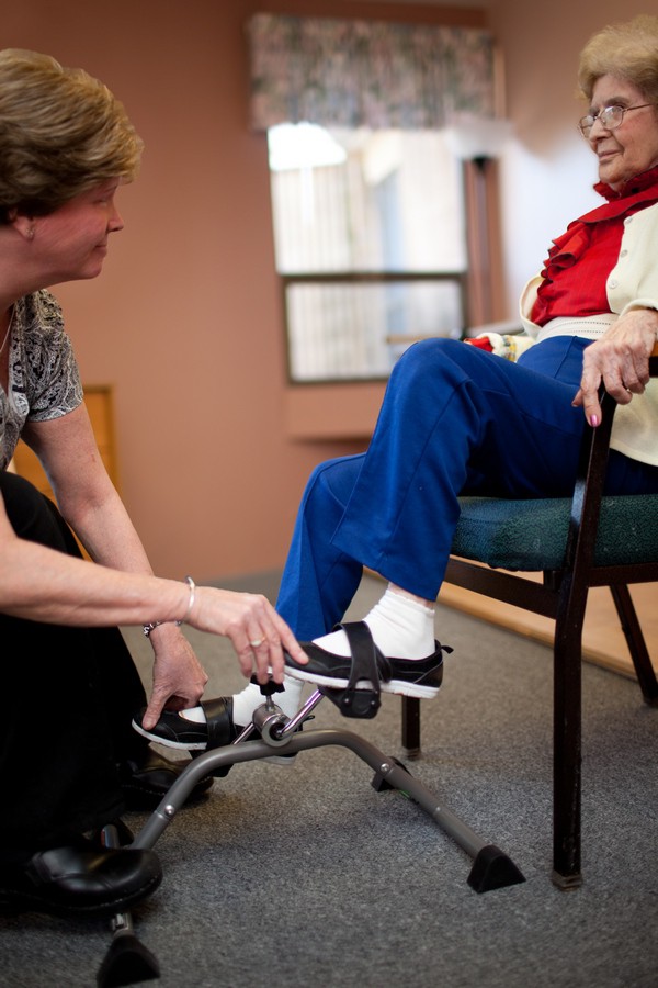 A caregiver assists an elderly person with a pedal exerciser for physical therapy.