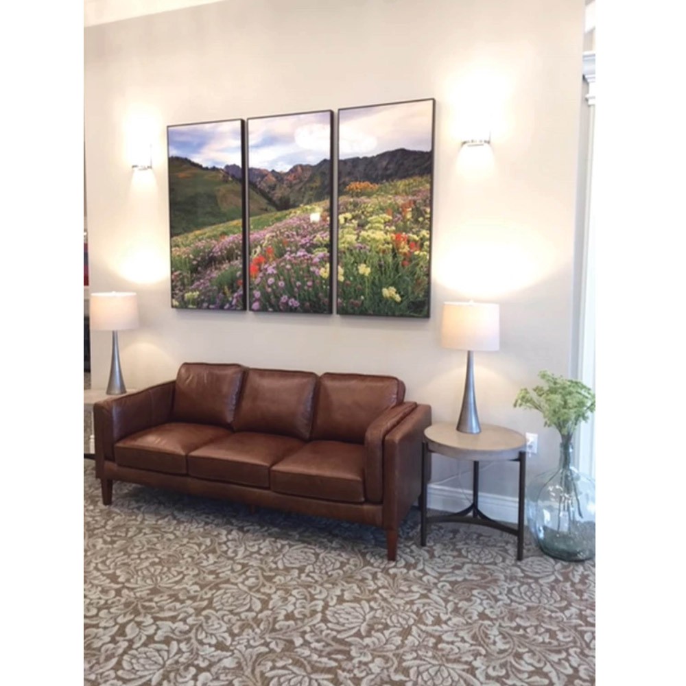 A cozy living room setup featuring a brown leather sofa, modern lamps, and a three-panel landscape painting.