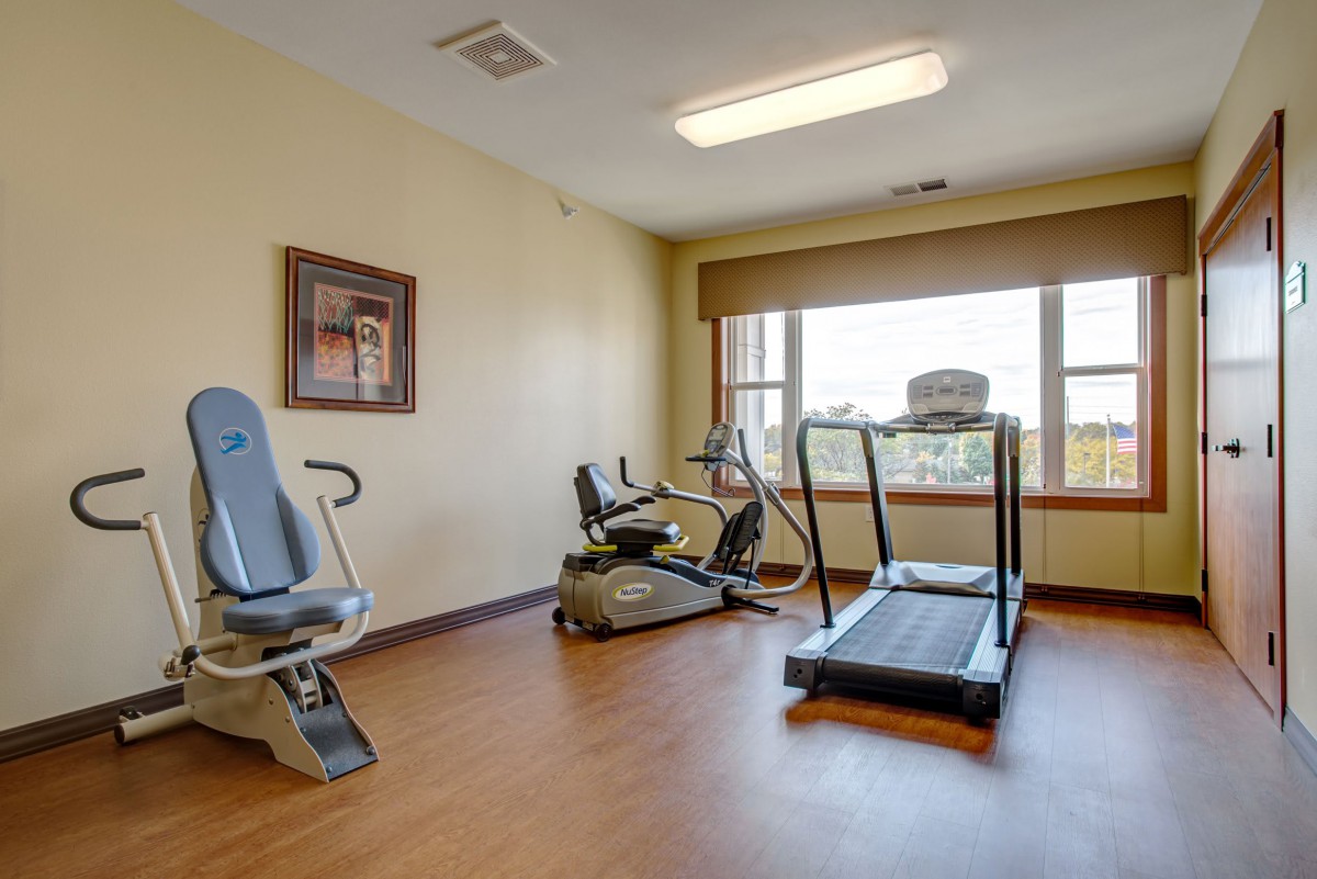 A small gym with fitness equipment, including a treadmill and stationary bike, located in a well-lit room with a large window.